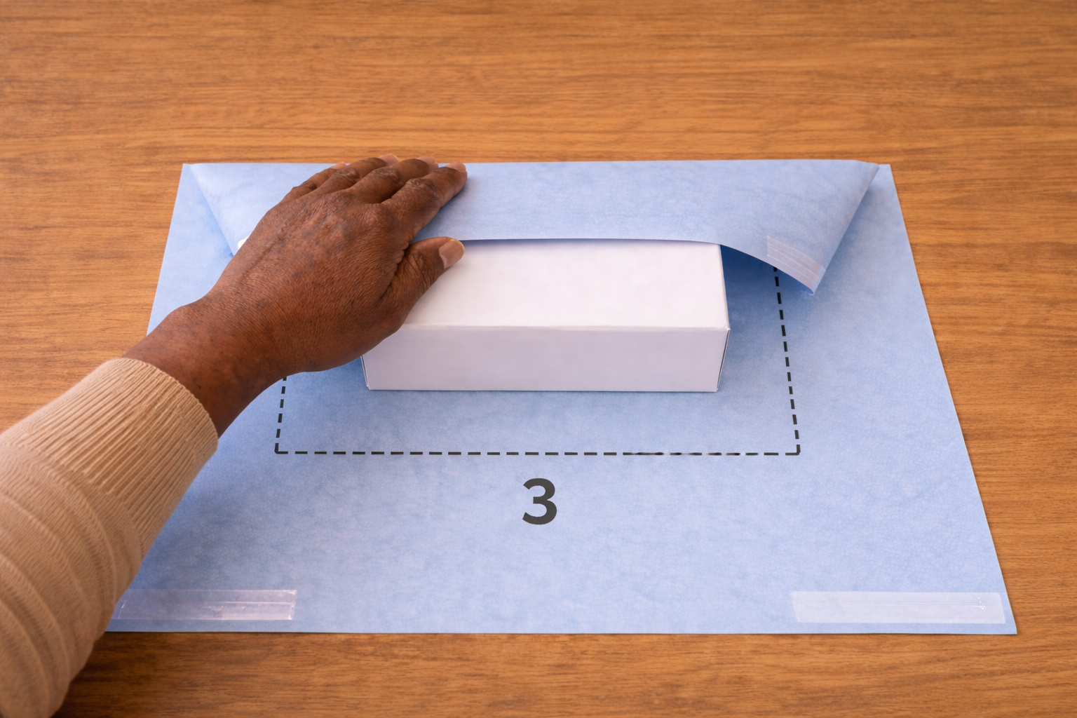 Hand folding number and dotted line guided blue wrapping paper over a white box on a wooden surface